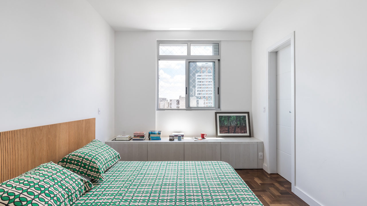 A suíte segue a mesma lógica estética e funcional do restante do apartamento, com pouco mobiliário para protagonizar o espaço. Sob a janela, o banco-gaveteiro organiza as peças da moradora. 